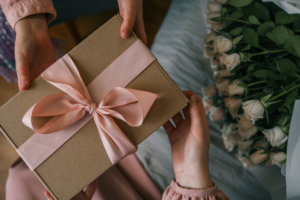 Hands of a child giving a wrapped gift with a satin ribbon to a woman holding a bouquet of roses, captured in warm indoor light. mother's day