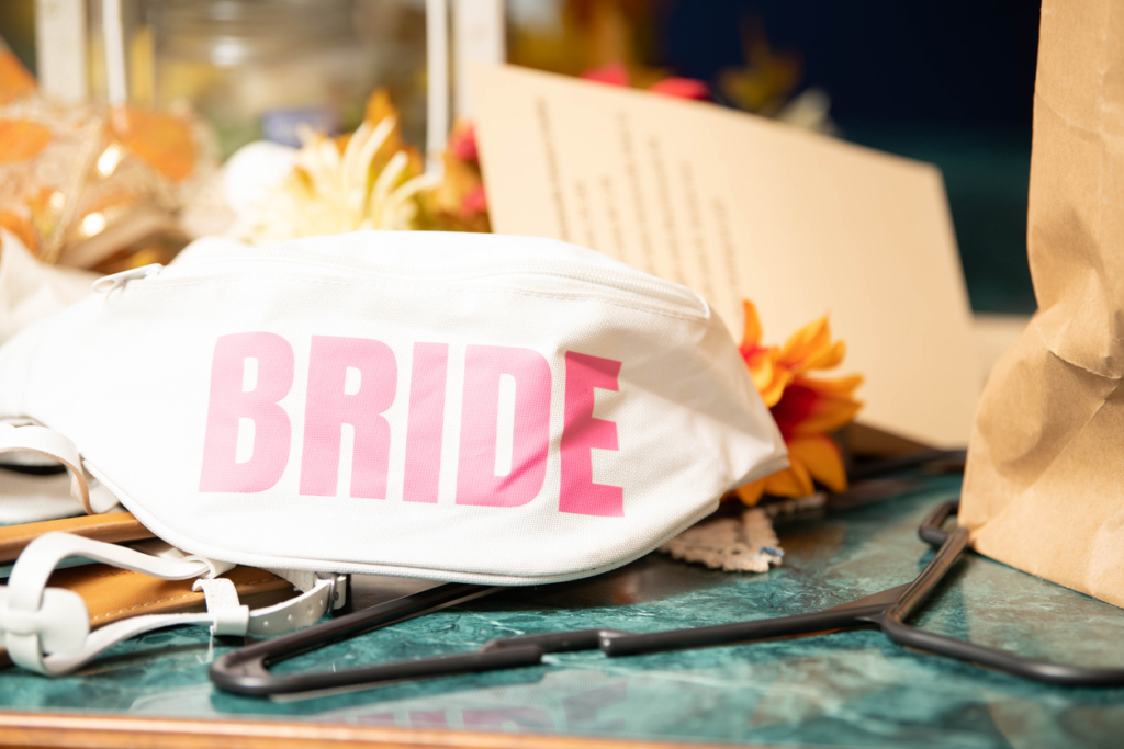 Trendy white fanny pack featuring bold pink "Bride" lettering worn by woman celebrating wedding. Shows contemporary bridal accessory combining function with style. Symbolizes modern bride breaking traditions, practical elegance, hands-free to embrace life, comfort in celebration, rejecting constraints, new generation's wedding style, and confidence to be yourself. Represents evolution of bridal fashion, accessibility meets beauty, casual sophistication, freedom of movement, authenticity over formality, and rewriting wedding rules. Metaphor for modern relationships, comfortable commitment, and being unbound by tradition. Perfect for illustrating modern weddings, bride trends, casual elegance, breaking tradition, contemporary brides, practical fashion, and authentic celebration. wedding day emergency kit essentials