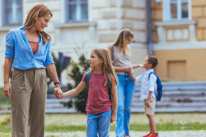 first day of school outfit ideas for moms A cheerful Caucasian woman in casual attire walks her young daughter to school, holding hands as they navigate a busy outdoor setting with other students around.