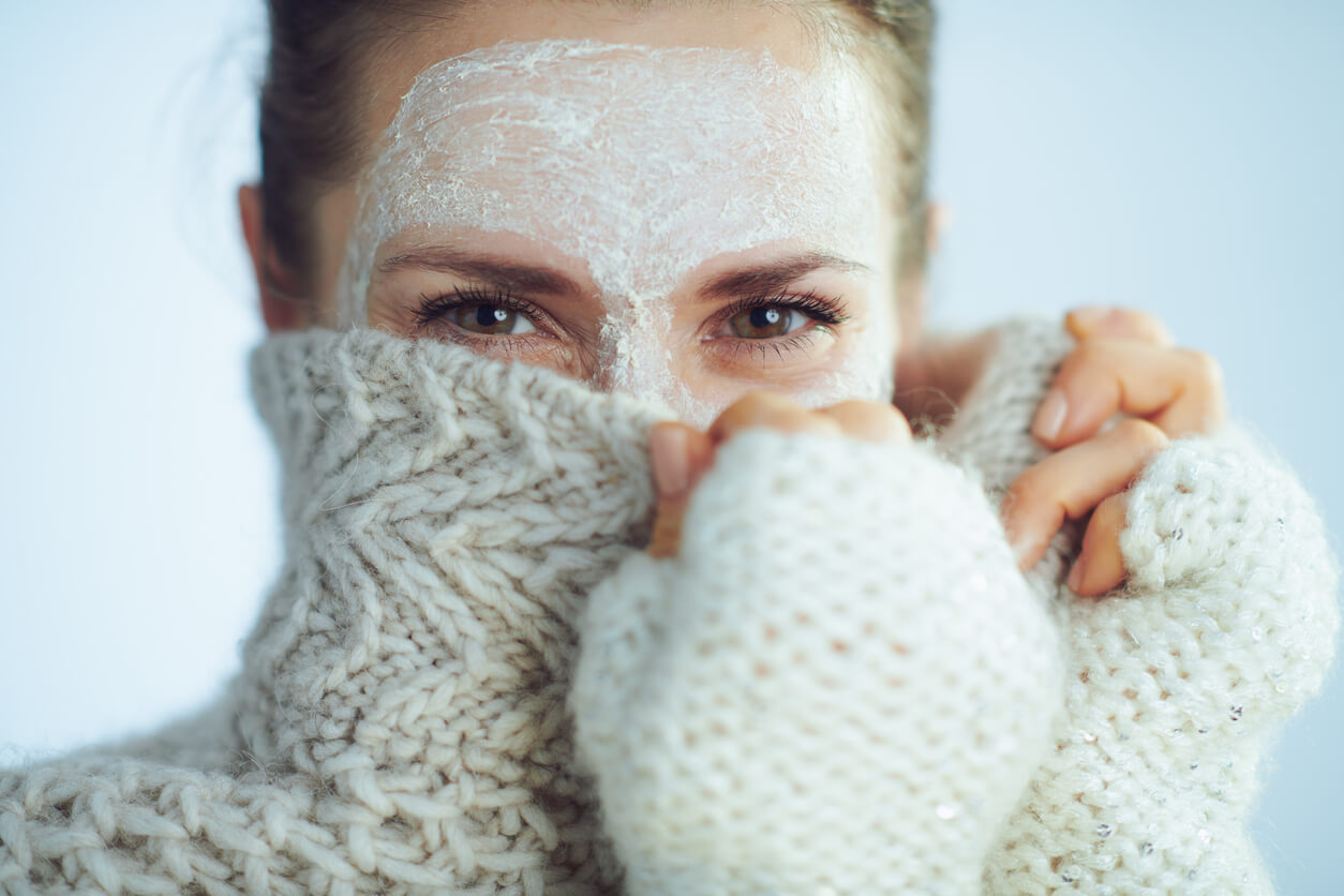skincare essentials skincare essential winter season woman with a face mask on in the winter elegant woman in roll neck sweater and cardigan with white facial mask hiding behind clothes on winter light blue background.