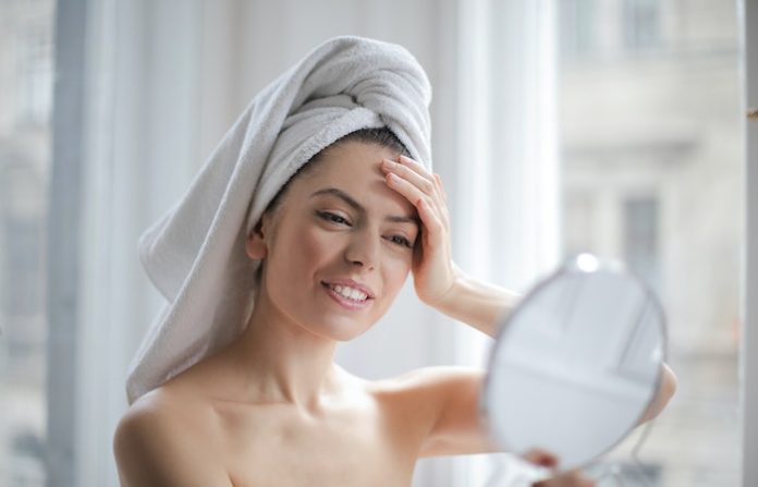 Selective Focus Portrait Photo of Smiling Woman With a Towel on Head Looking in the Mirror - How to Improve Skincare Routine