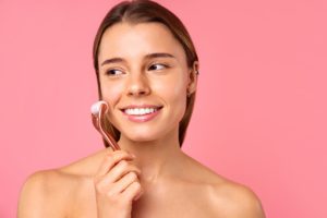 Beautiful woman doing massage for face lift and anti wrinkle using microneedling mesotherapy and serum isolated on pink background. Facial skin care, cosmetology, beauty concept. ice rolling