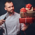 Chef holding gifts and spatula.Portrait of bearded chef cook holds colorful paper Christmas gifts on grey background. gifts for chefs