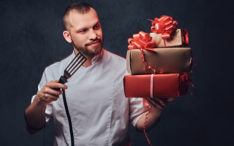 Chef holding gifts and spatula.Portrait of bearded chef cook holds colorful paper Christmas gifts on grey background. gifts for chefs