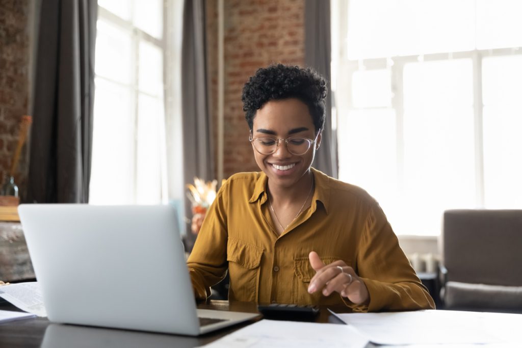 Happy young Afro American entrepreneur woman in glasses counting profit, on calculator at laptop computer, analyzing benefits, enjoying financial success, job high result, smiling file taxes jointly or separately 2026