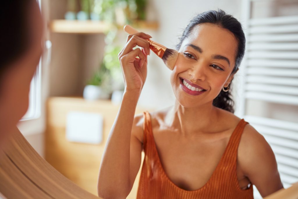 woman doing her makeup spring break beauty routine Beautiful girl applying makeup using powder brush before going to work. Healthy latin woman looking in the mirror and applying cosmetic with a big brush. Young woman looking in the mirror and applying foundation or blusher on her face.