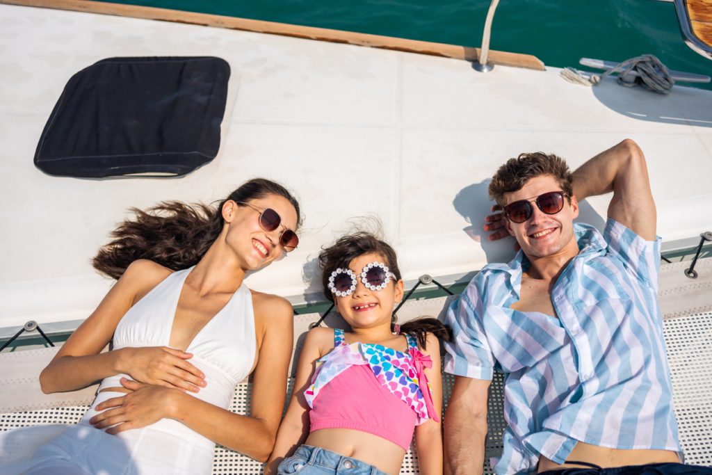 affordable summer travel ideas families Caucasian attractive family lying down on deck of yacht while yachting. Young beautiful couple hanging out and spend time with daughter while catamaran boat sailing during holiday summer trip together