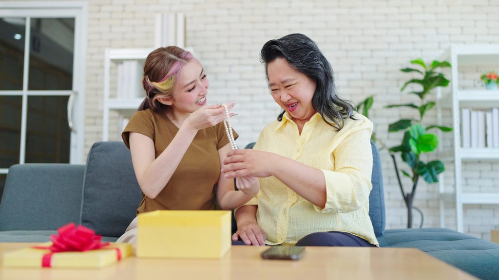 Young Asian daughter surprised gifting birthday present to mother while sitting on sofa at home. Young woman giving birthday gift to mother. Happy family concept, Mother's day concept mother's day jewelry gifts